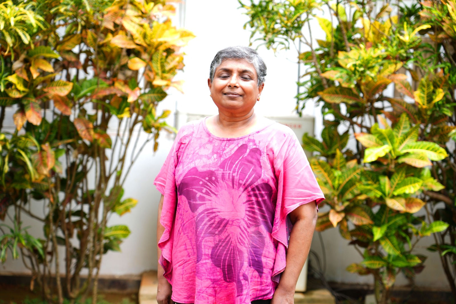 Shalika Karunaratne is standing in front of a white wall, surrounded by green foliage. She has short grey hair and wears a vibrant pink Tshirt which has a large floral design